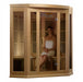 Woman sitting inside a wooden infrared sauna with glass panels.
