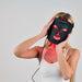 Woman using a black LED face mask with red lights on a plain background