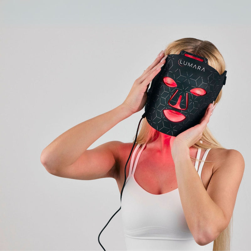 Woman using a black LED face mask with red lights on a plain background